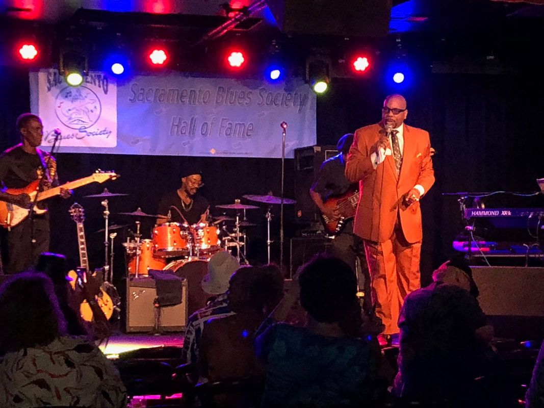 Live band performing on a small stage under red and blue lights, with singer in orange suit center stage.