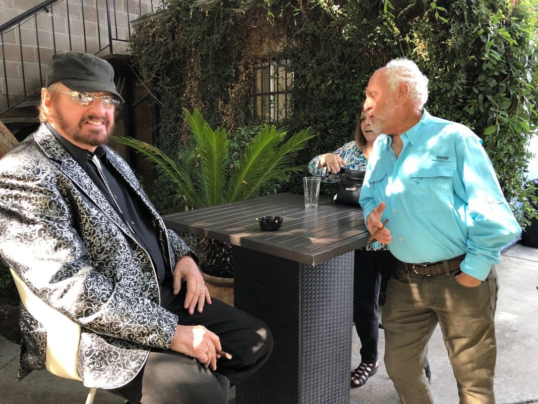 Two people talking at an outdoor table in a garden patio, one seated and one standing with a drink.
