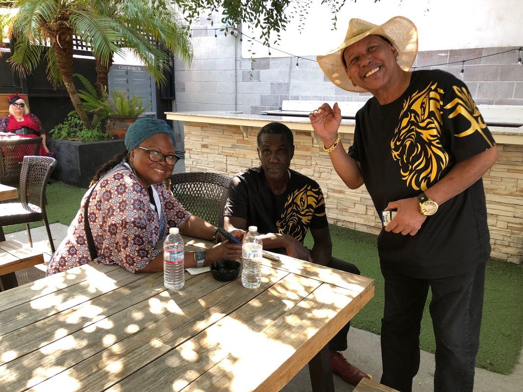 Three people sitting and standing around a table outdoors, smiling in the shade near a building.