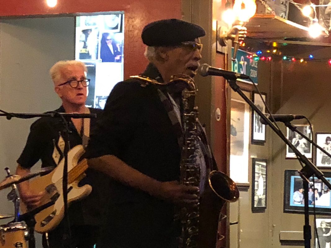 Two musicians perform indoors, one singing into a microphone while another plays bass behind him.