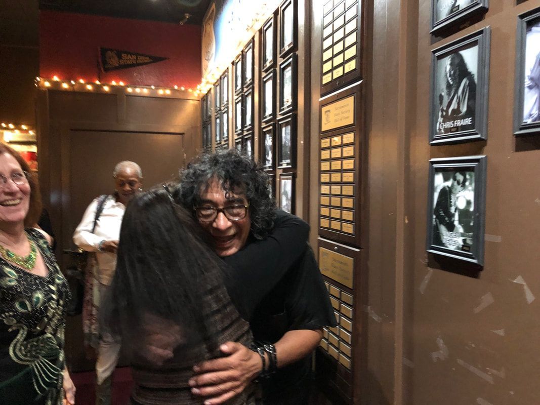 Two people hug in a warmly lit hallway lined with framed photos.