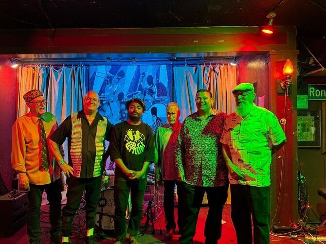Six people standing on a small stage under red and green lights, with a blue curtain backdrop