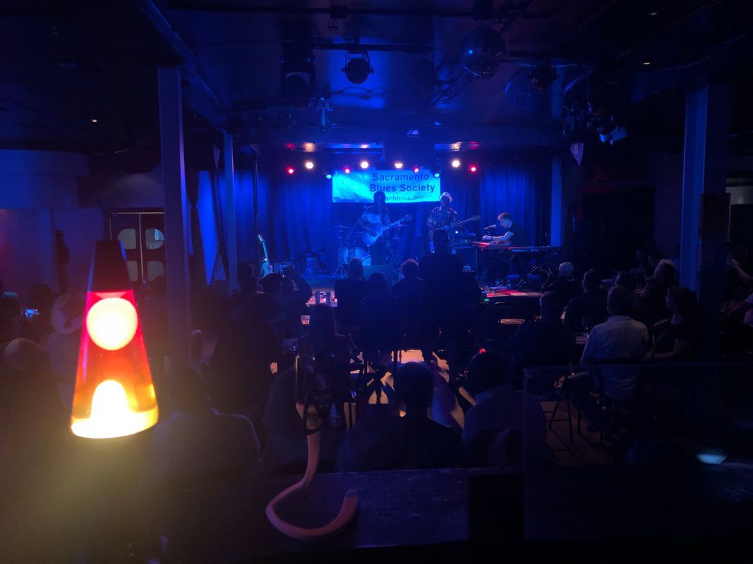 Dim nightclub or bar with blue stage lights and a glowing lava lamp in the foreground.