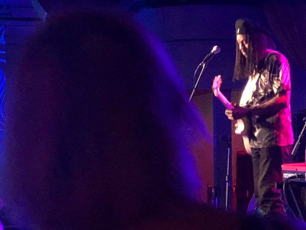 Guitarist performing on a purple-lit stage, seen from the crowd with a microphone stand nearby