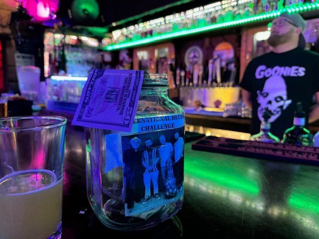 Dim neon bar counter with a glass tipped by cash, lit in purple and green.