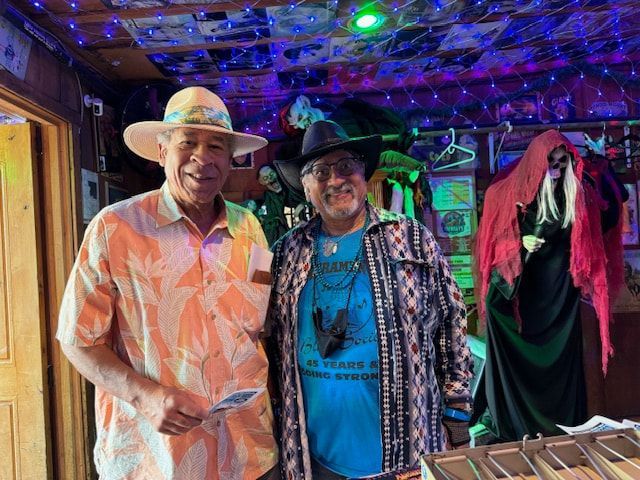Two people smiling indoors under colorful string lights, wearing hats and patterned shirts