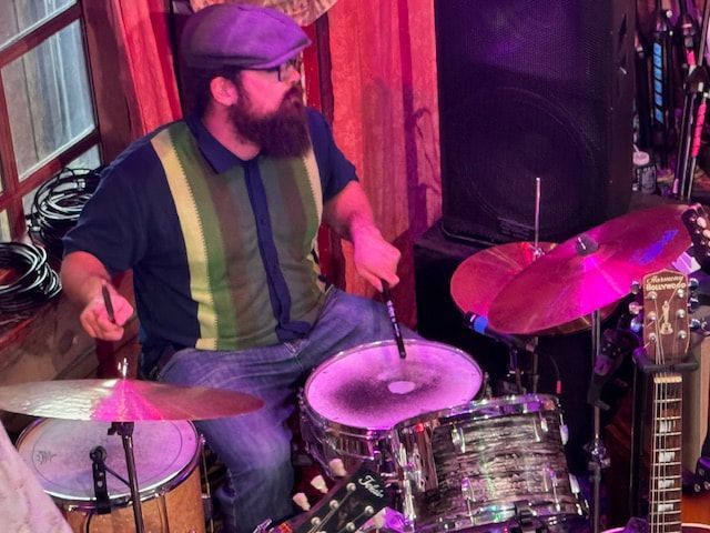 Drummer in a green striped shirt playing drums beside a guitar and speaker on a small stage