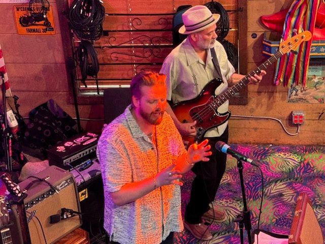Musicians performing in a colorful room, one singing into a mic while another plays bass guitar.