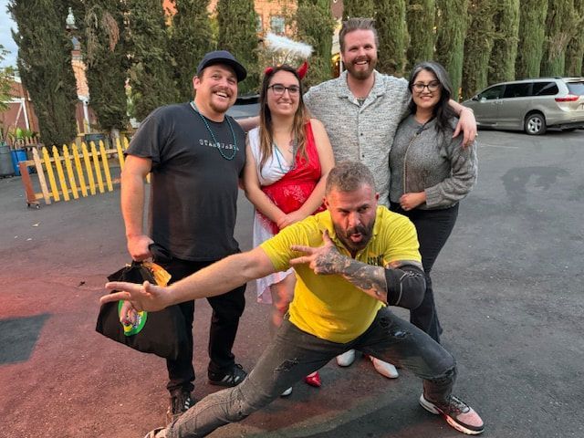 Five people posing outdoors, one crouching in front with arms outstretched, others smiling behind him.