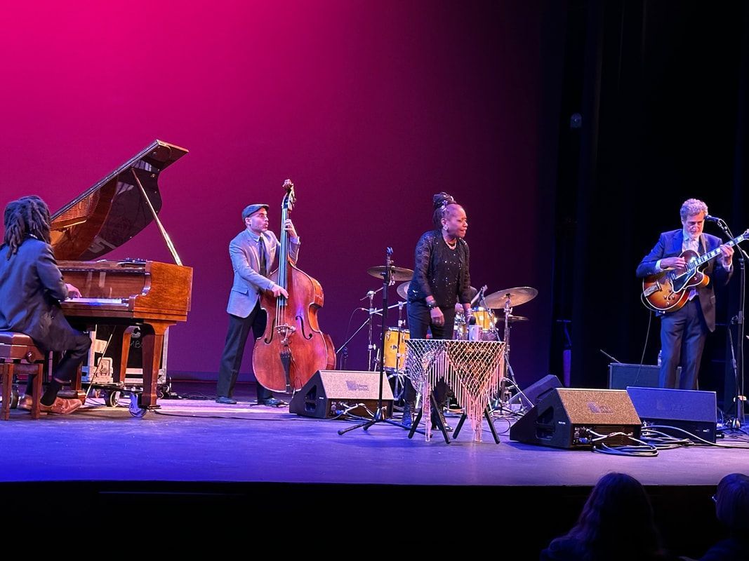 Jazz band performing onstage under pink and blue lights, with piano, bass, drums, and guitar.
