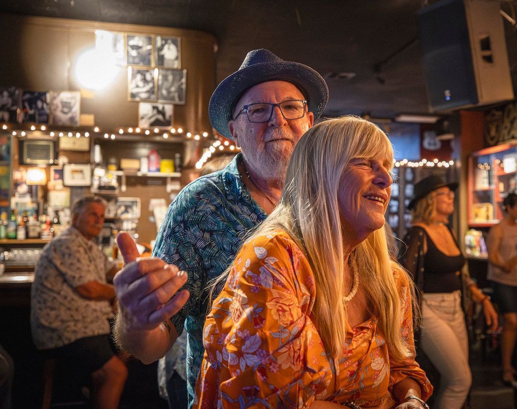 Two people smiling and dancing in a warmly lit bar, with shelves and patrons in the background.
