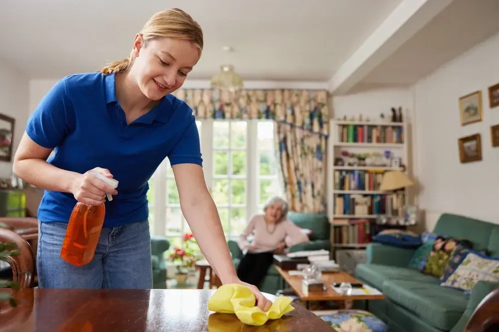 Person in a Blue Shirt Cleans a Wooden Table With a Spray Bottle — CQ Home Care in Yeppoon, QLD