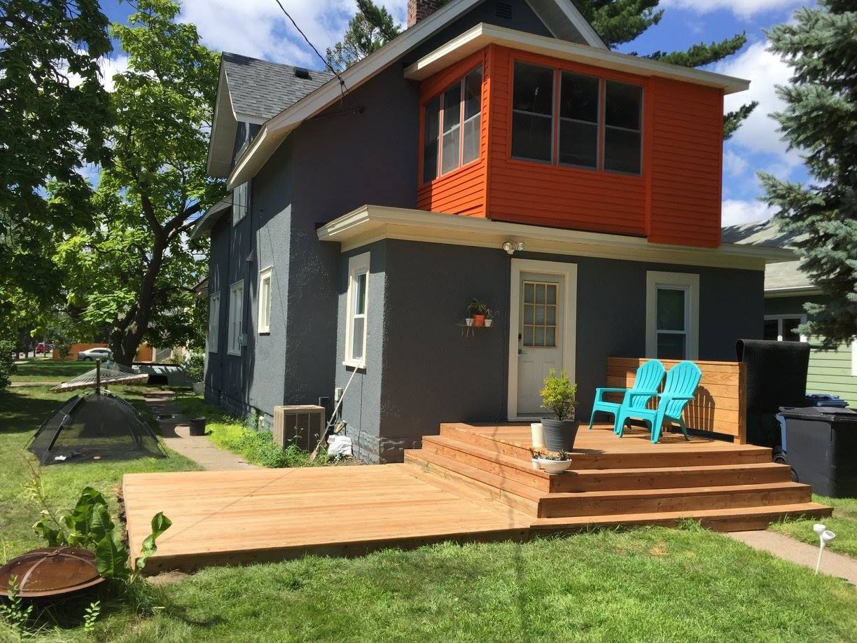 Two-story house with a wooden deck and orange accent, blue chairs.