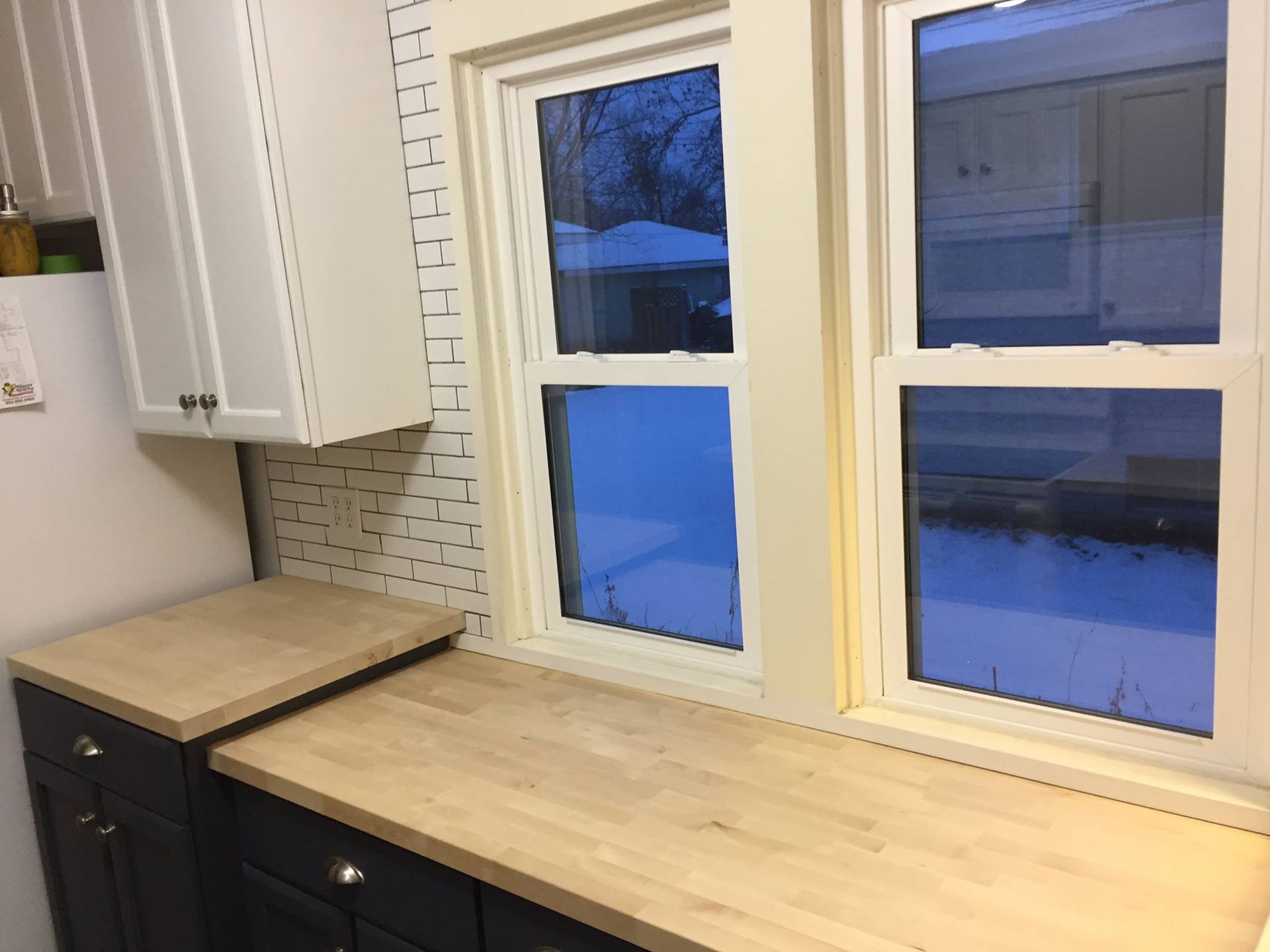 Kitchen with dark cabinets, light wood countertop, white brick backsplash, windows with winter view, and white upper cabinet.