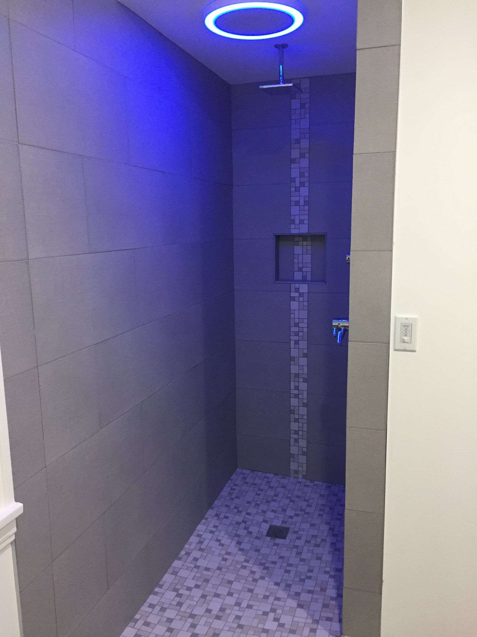 Modern shower with blue lighting. Gray tiled walls and pebble floor. Recessed shelf and circular ceiling light.