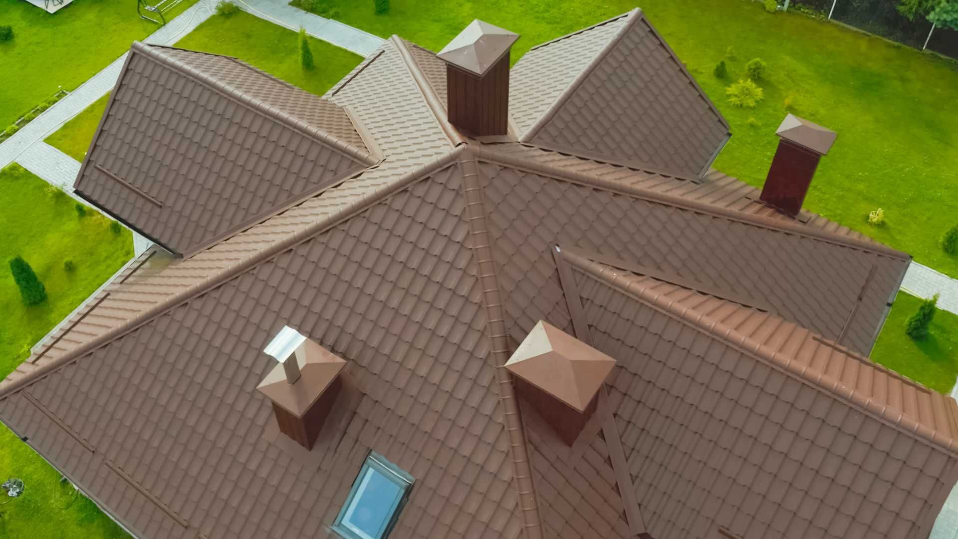 Brown roof of a house with multiple sections and chimneys, aerial view, set on green grass.