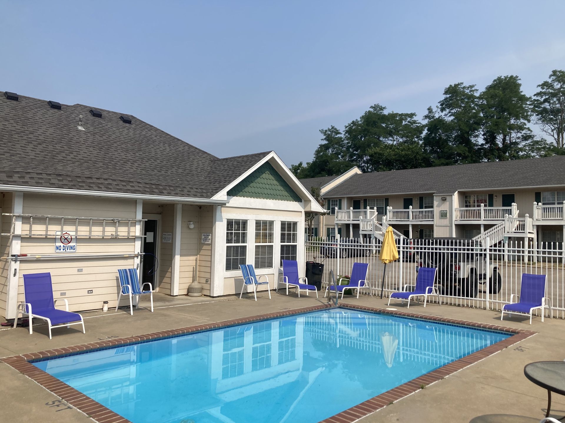Swimming pool with blue loungers and adjacent beige building with a dark roof.