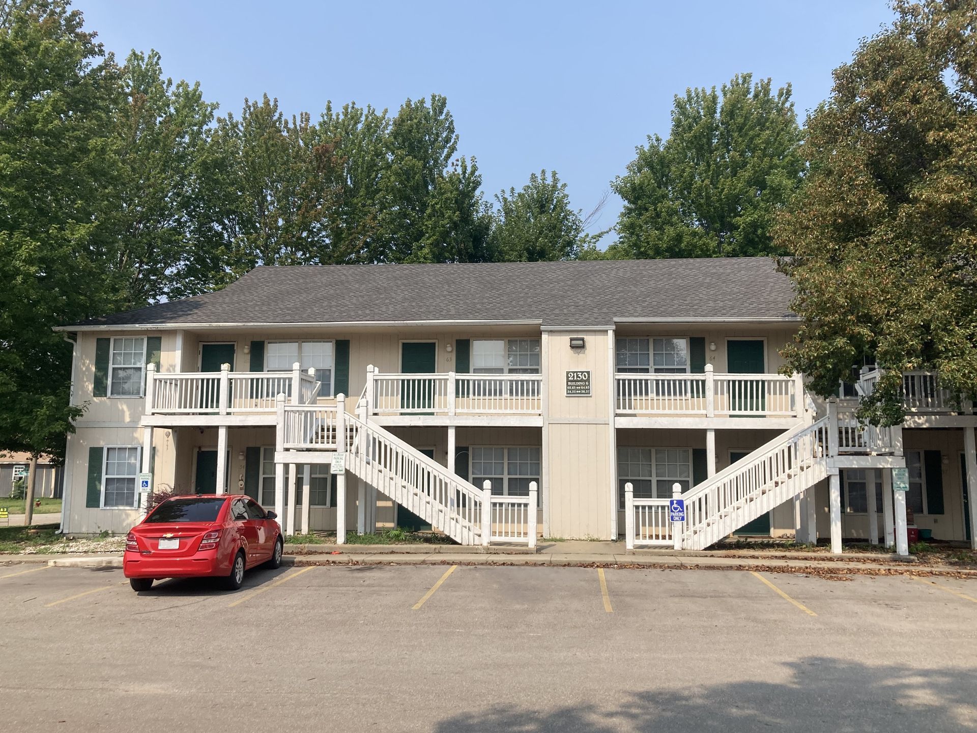 Two-story apartment building with white stairs and balconies. A red car is parked in front.