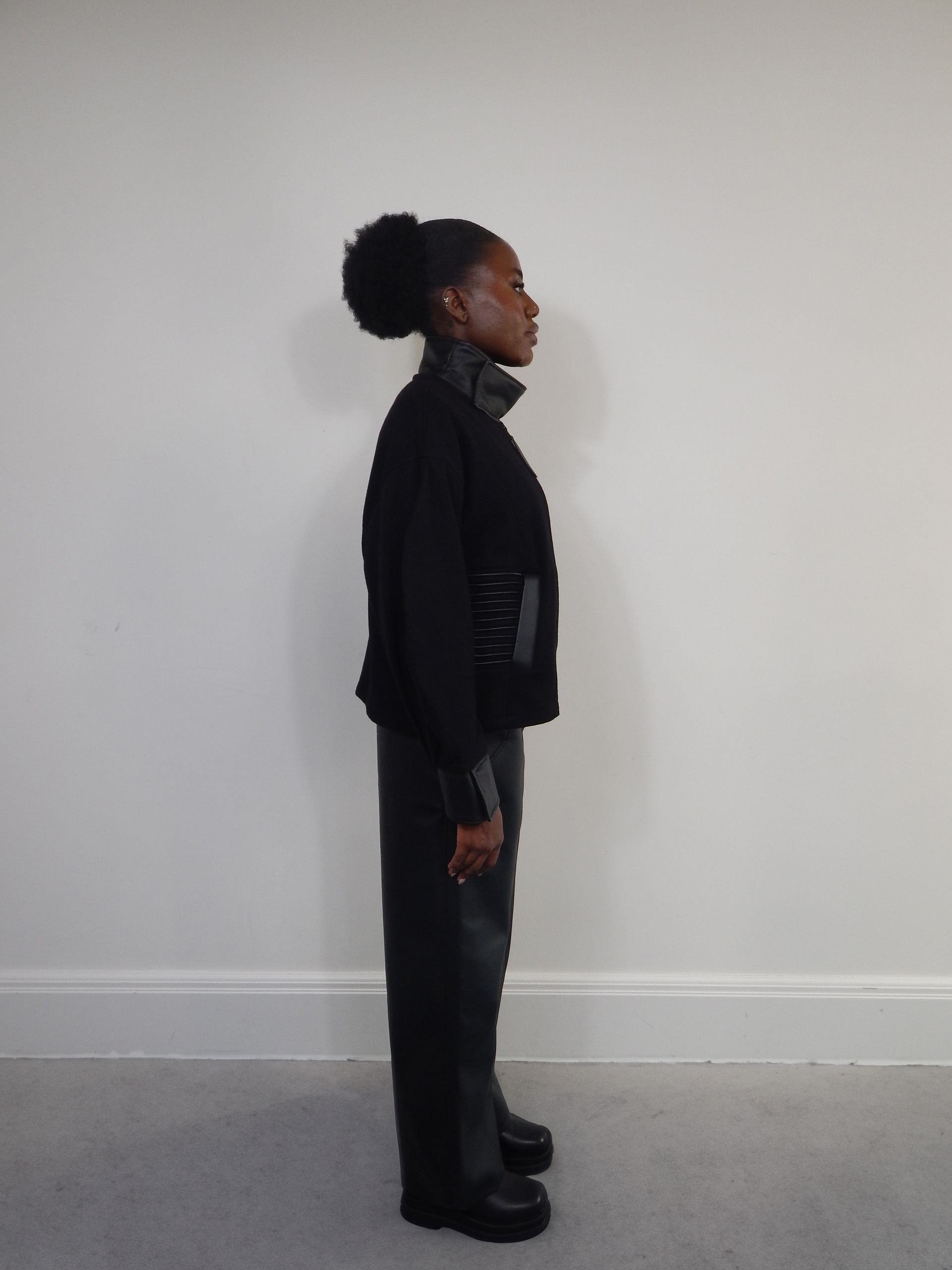 Person in black outfit standing in profile against a white wall.