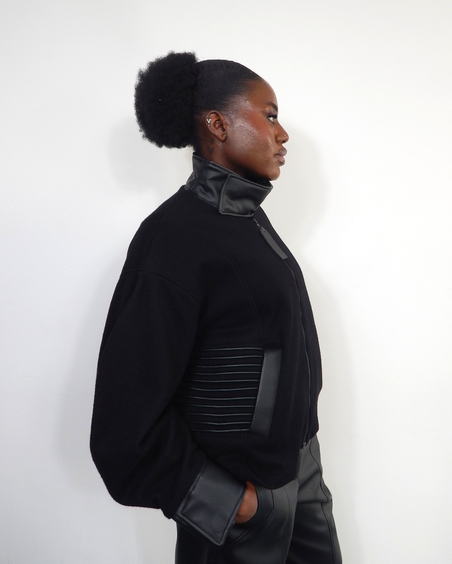 Woman in black jacket with leather details, hair up. White background.