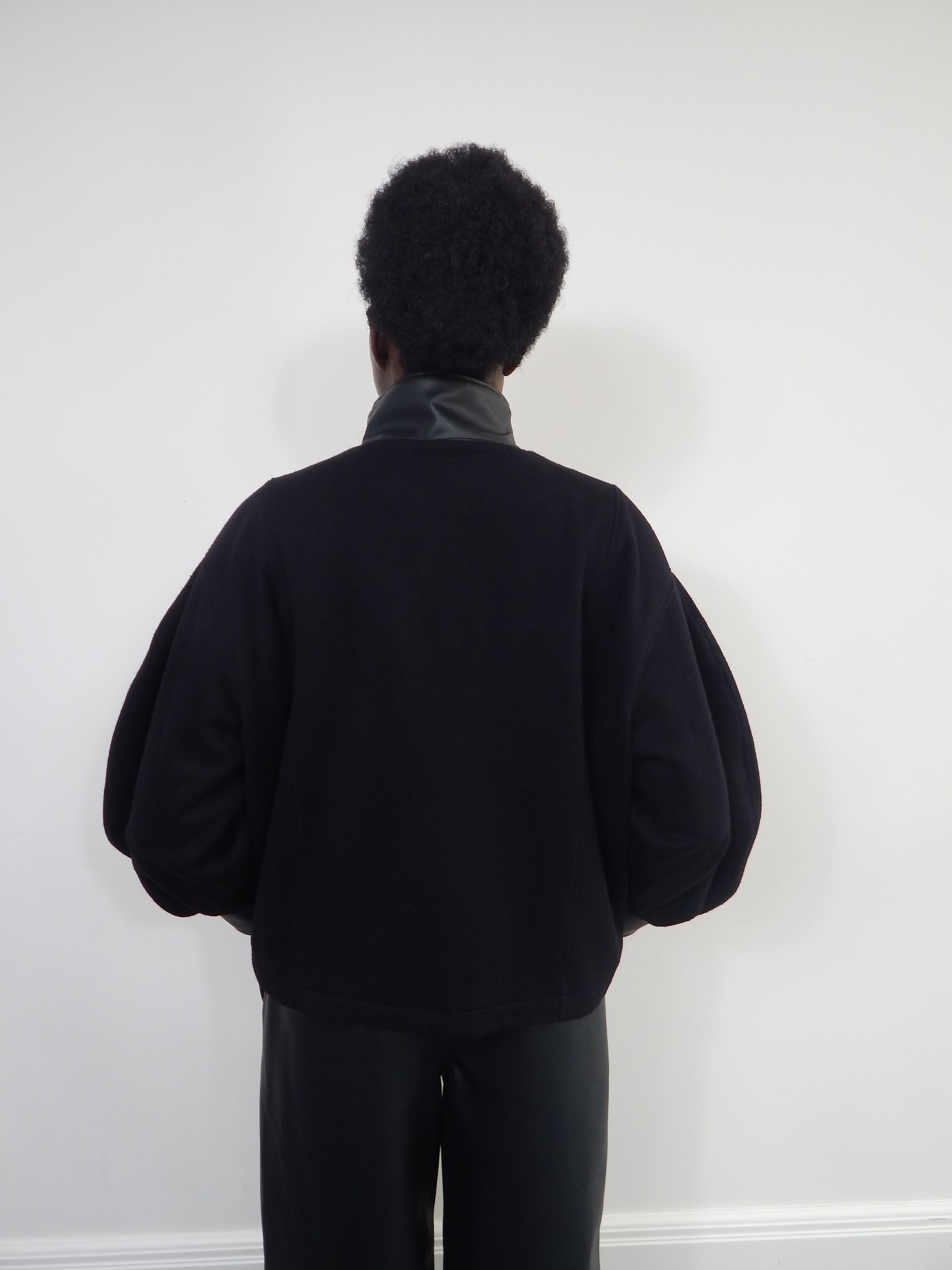 Back view of person in black sweater with puffy sleeves, standing against a white wall.