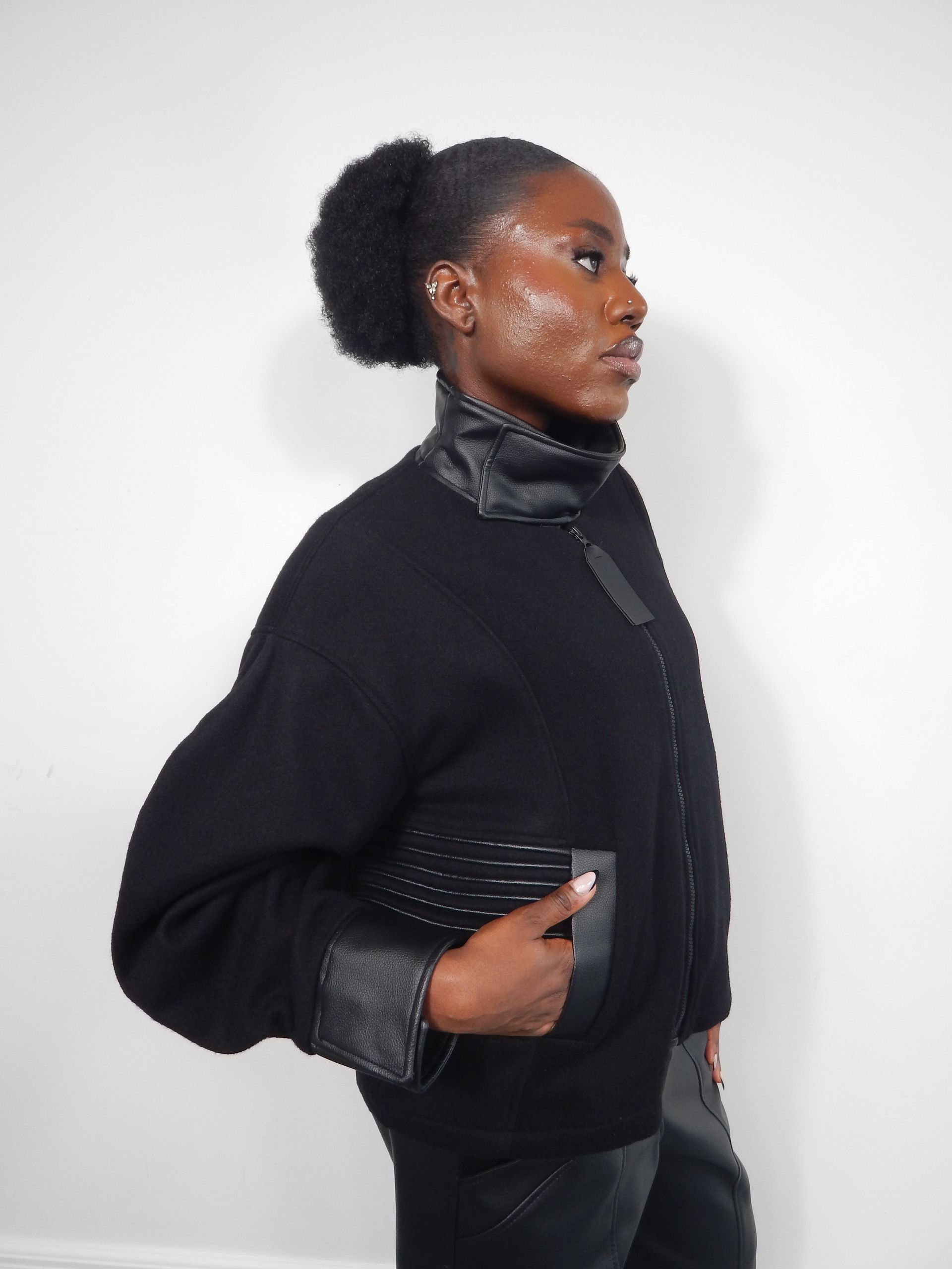Woman in black sweater with leather accents, holding a studded clutch, against a white wall.