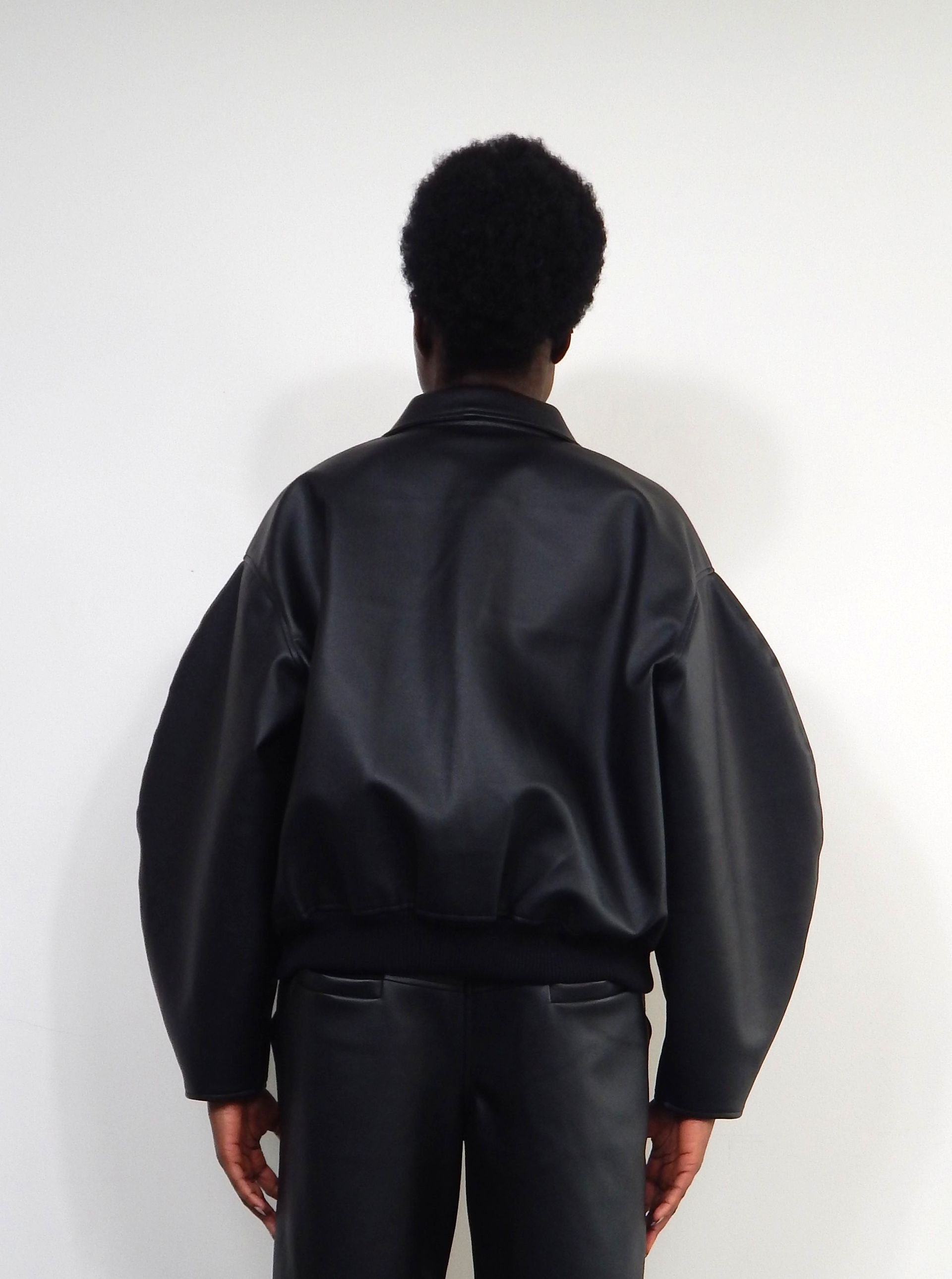 Person wearing a black bomber jacket with oversized sleeves, black pants, and standing in front of a white wall.