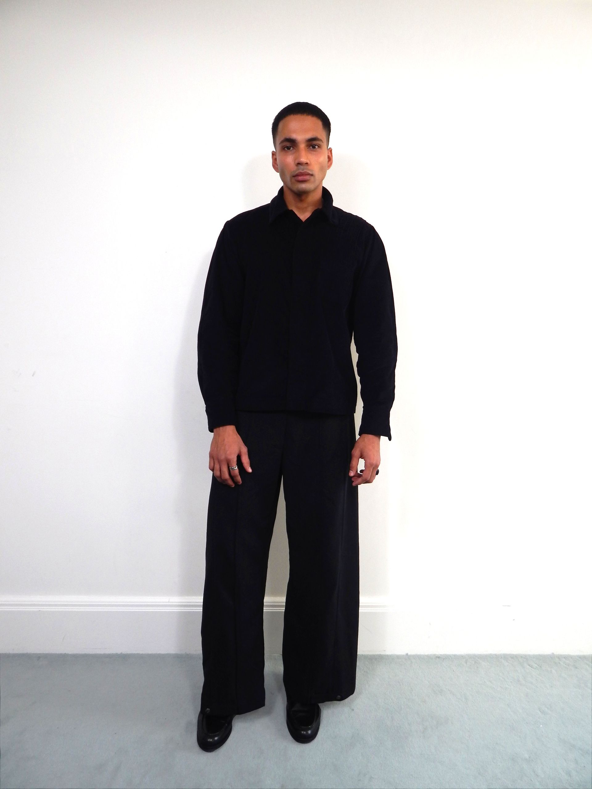 Person in black sweater and pants, standing against a white wall.