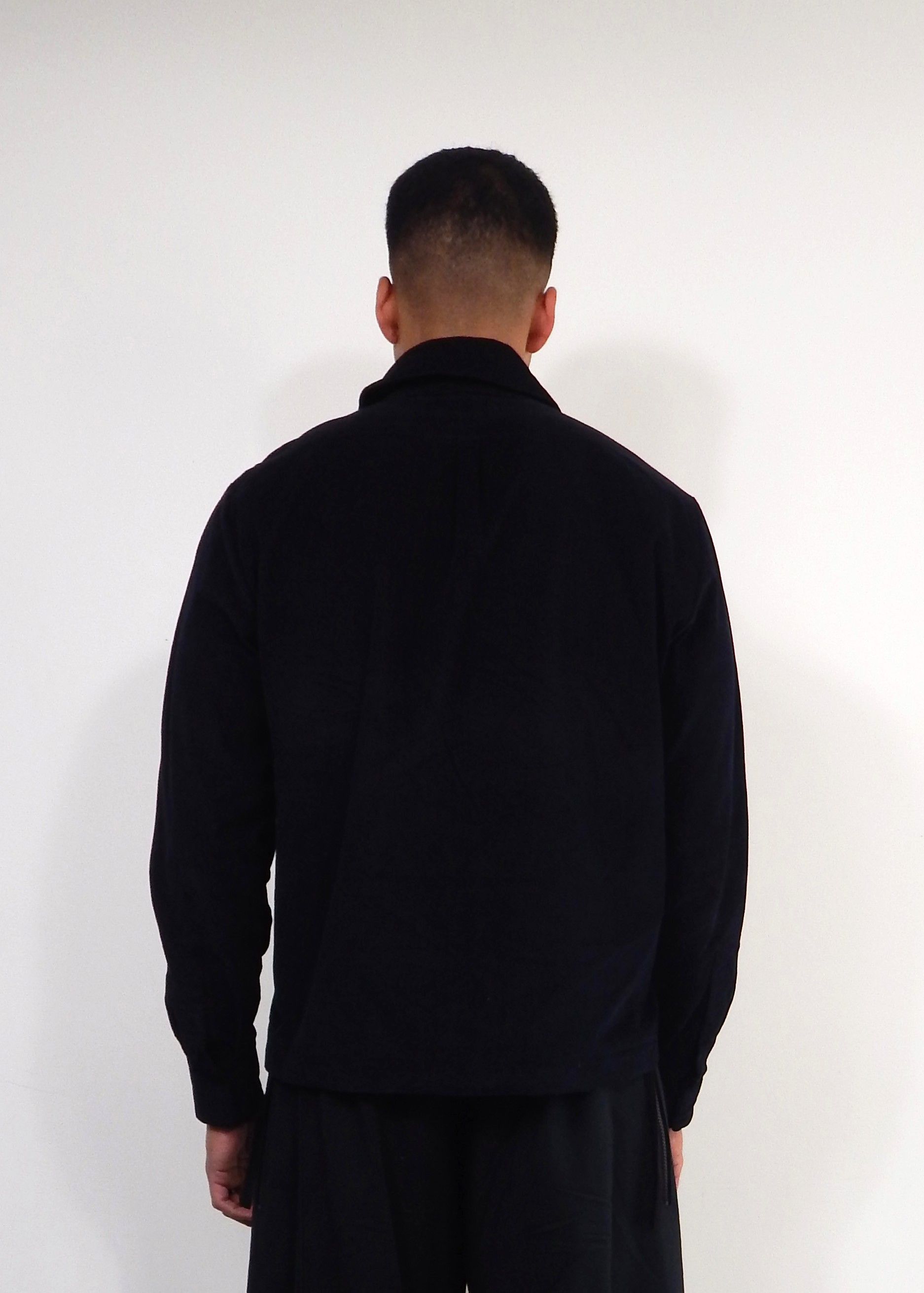 Person in a black jacket and pants, facing away from the camera, standing in front of a white wall.