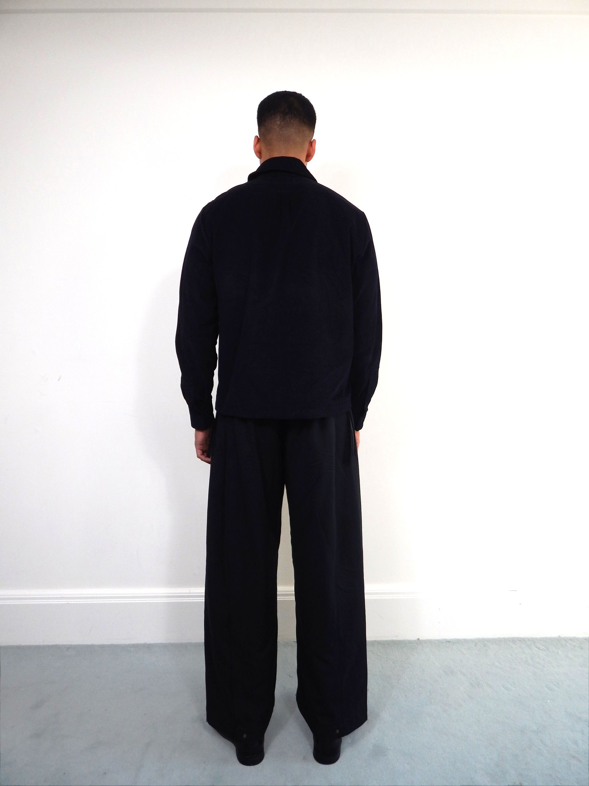 Person in dark clothing stands with back to the camera against a white wall.