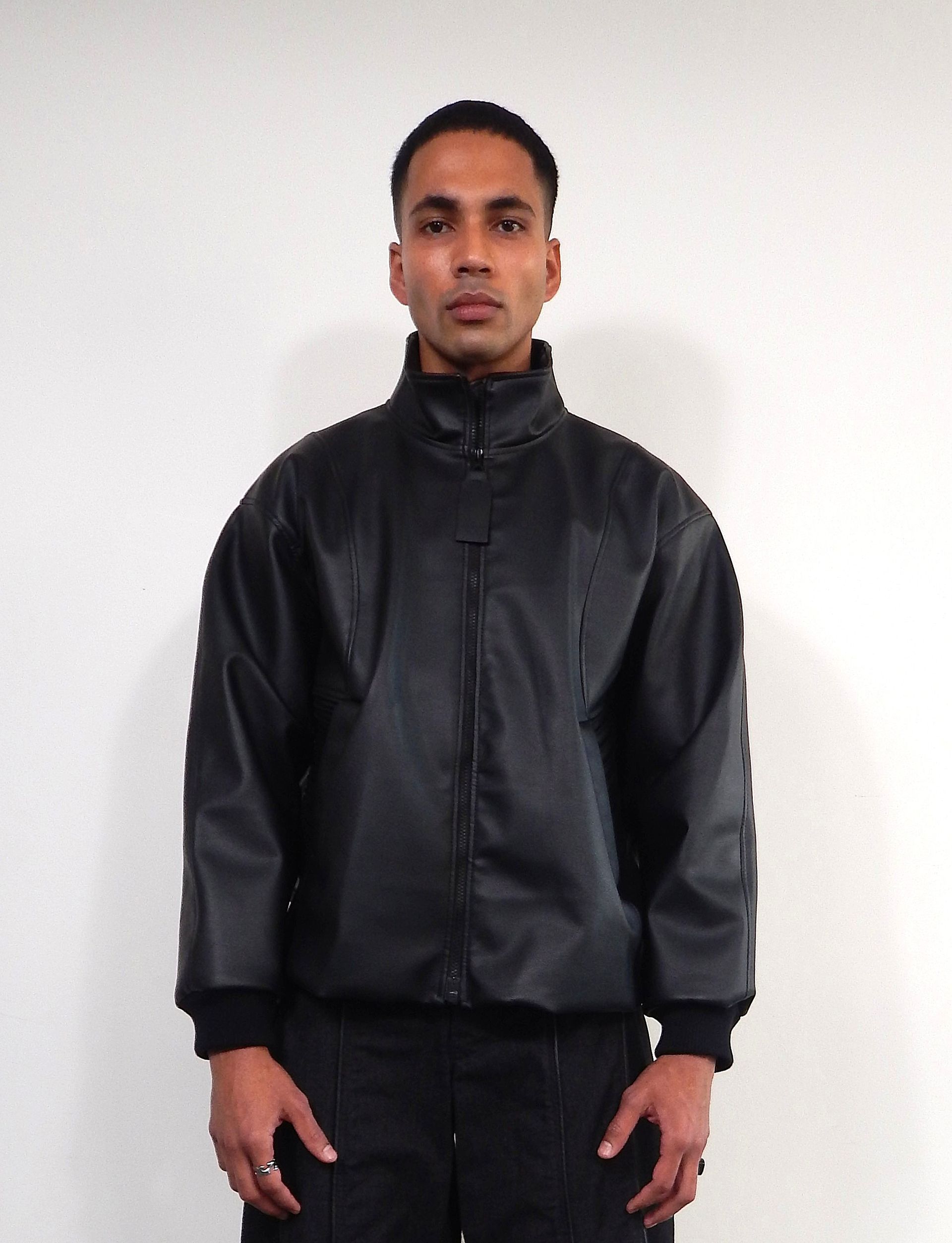 Man wearing a black jacket and black pants, standing in front of a white wall.