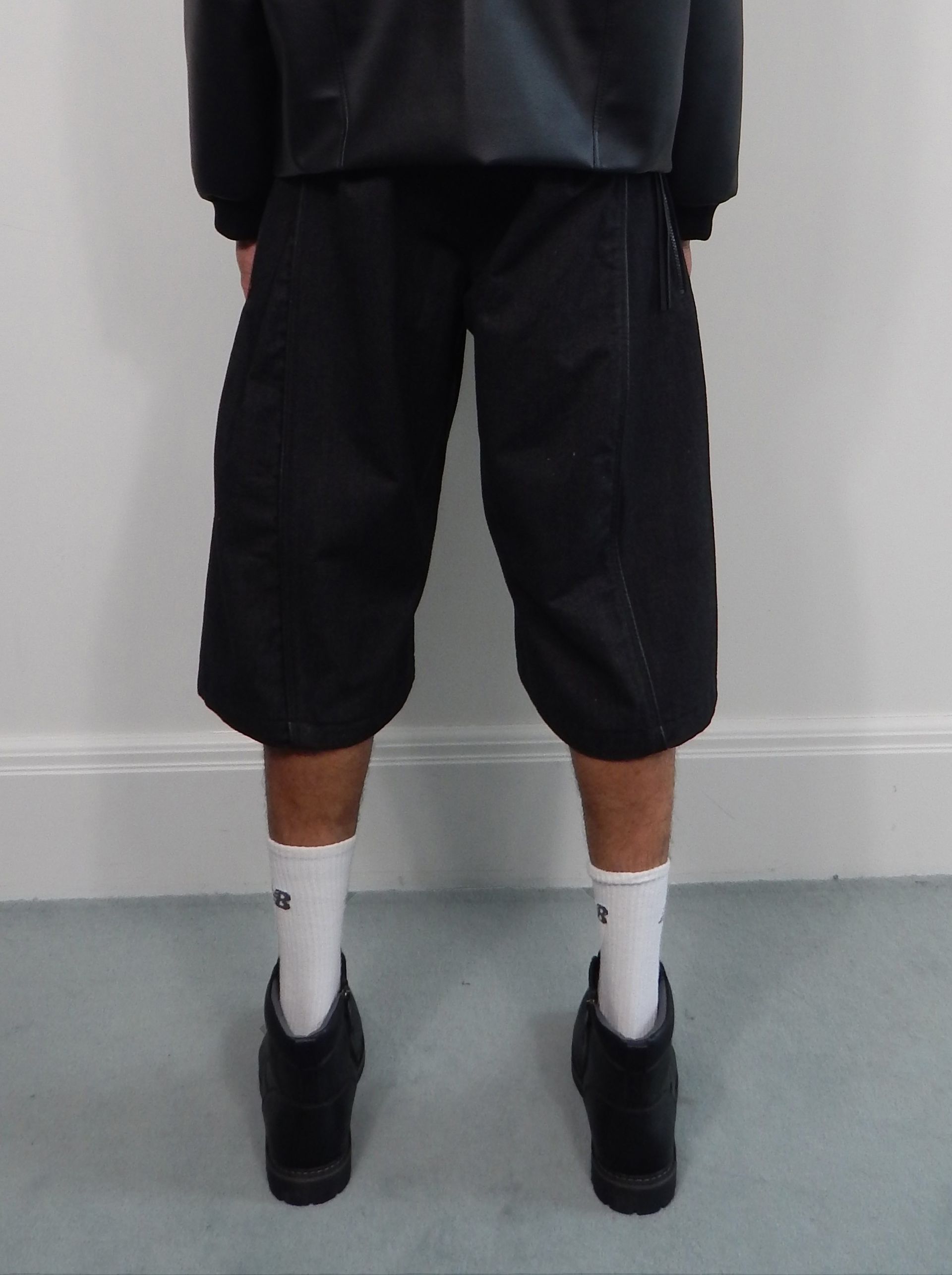 Person wearing black shorts, jacket, white socks, and black shoes, standing against a white wall.