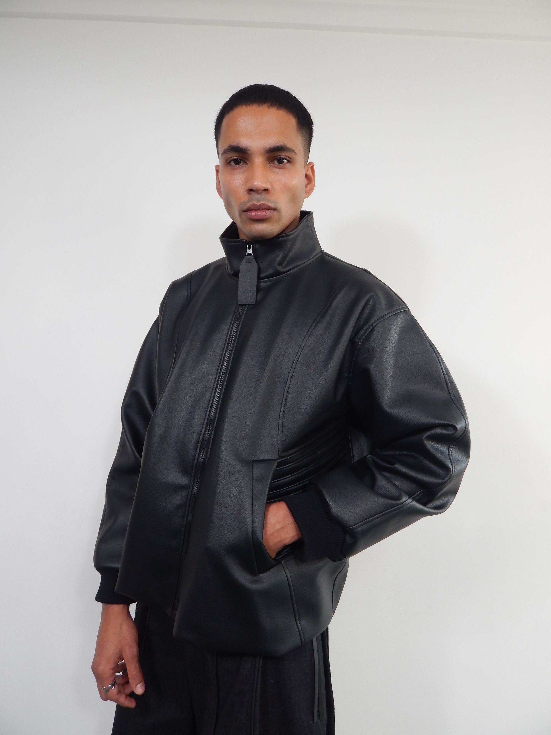 Person in black jacket with zipper and pockets, standing in front of a white wall.