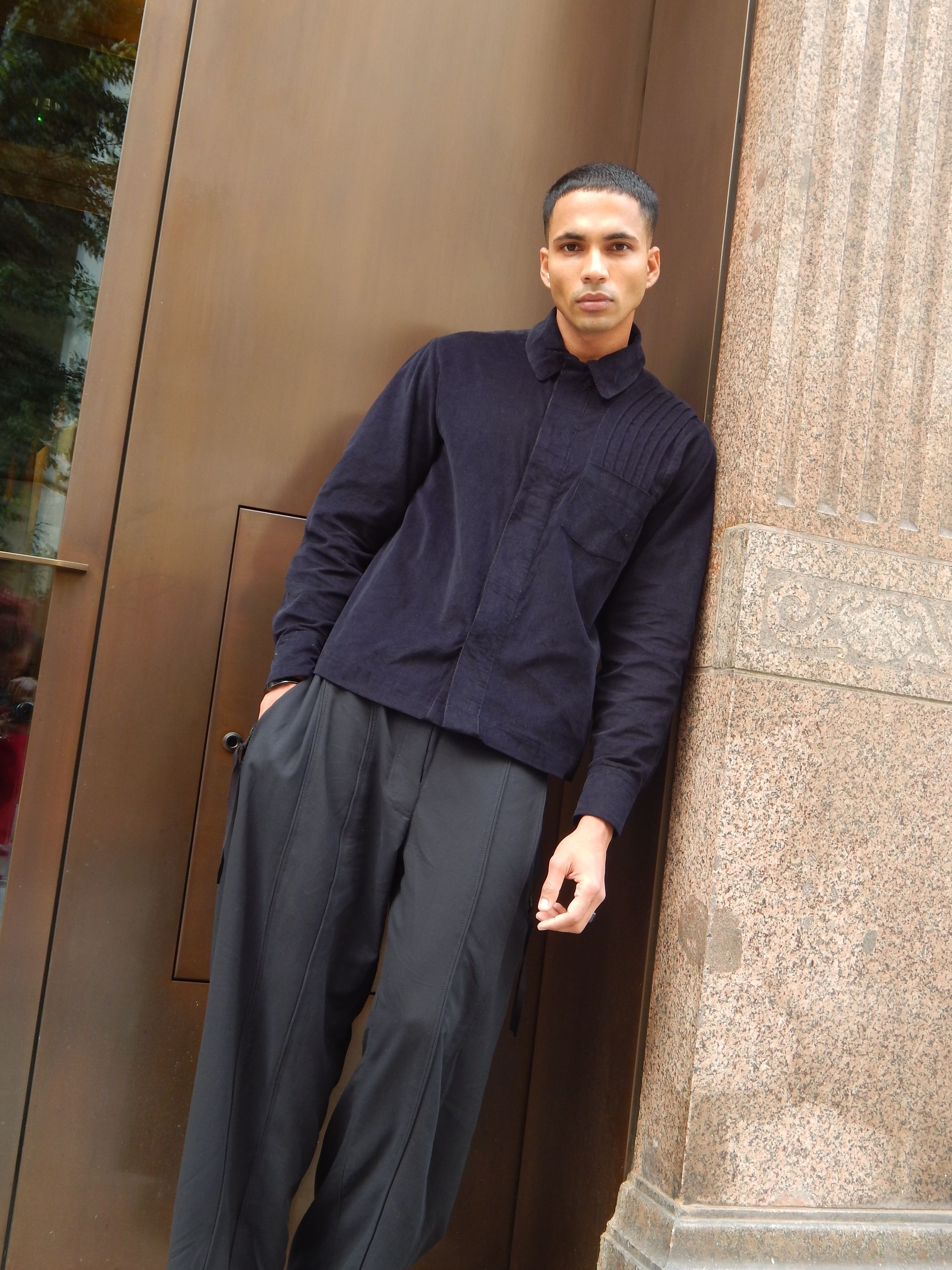 Man in navy shirt and black pants leans against a brown structure.
