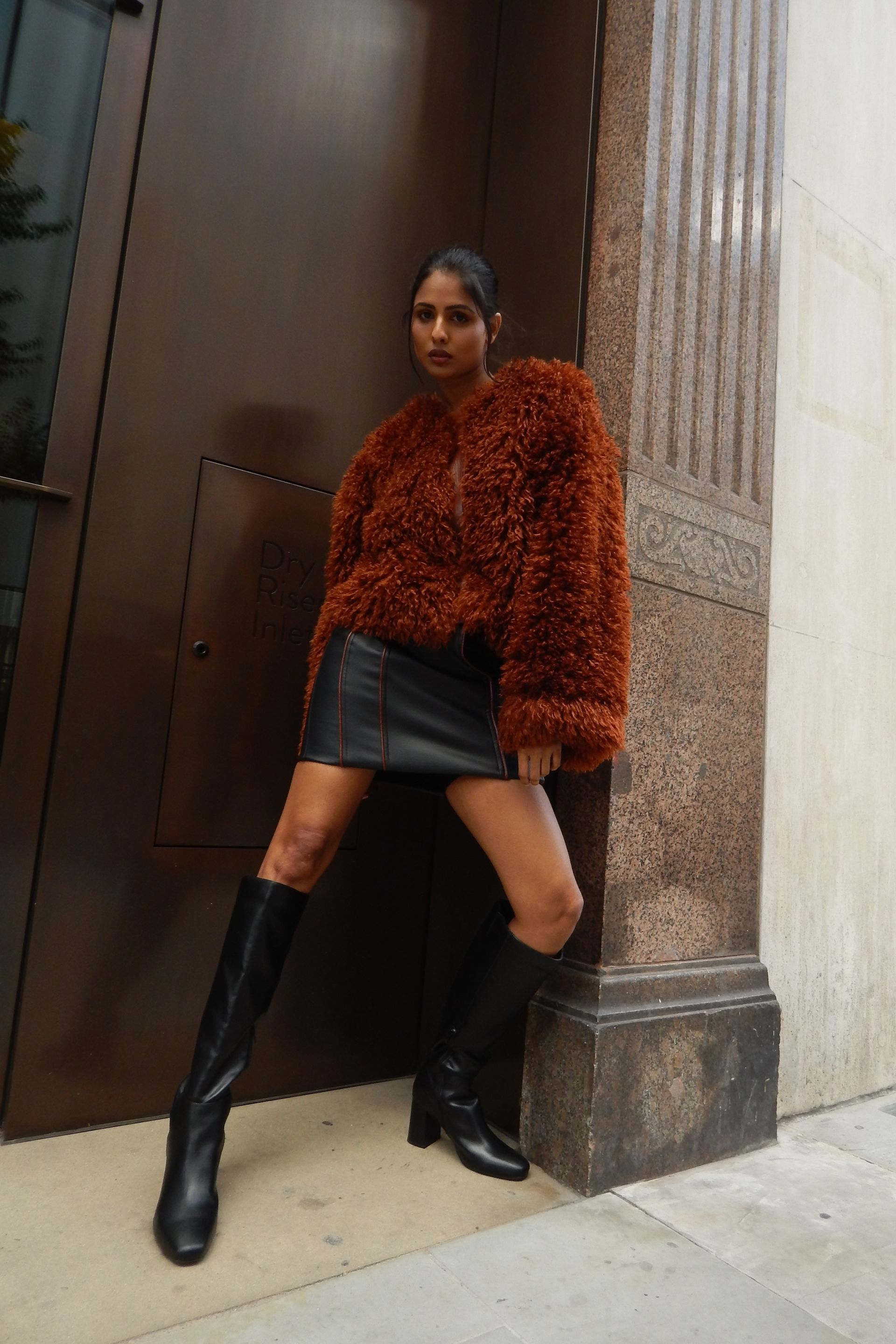 Woman in a fuzzy brown jacket, black skirt, and boots poses by a brown door and column.