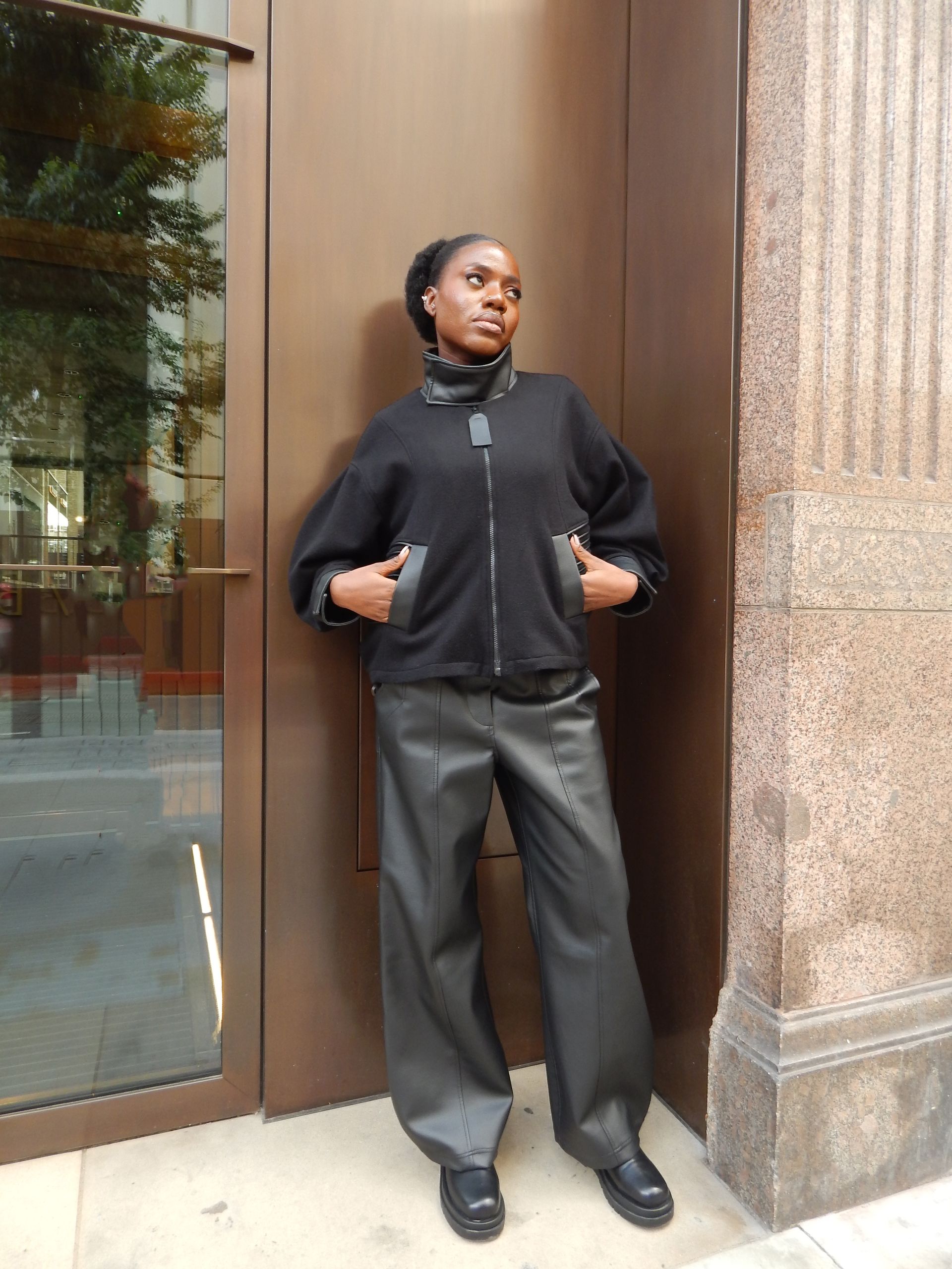 Person in black jacket and pants, hands in pockets, leaning against a brown door and textured wall.