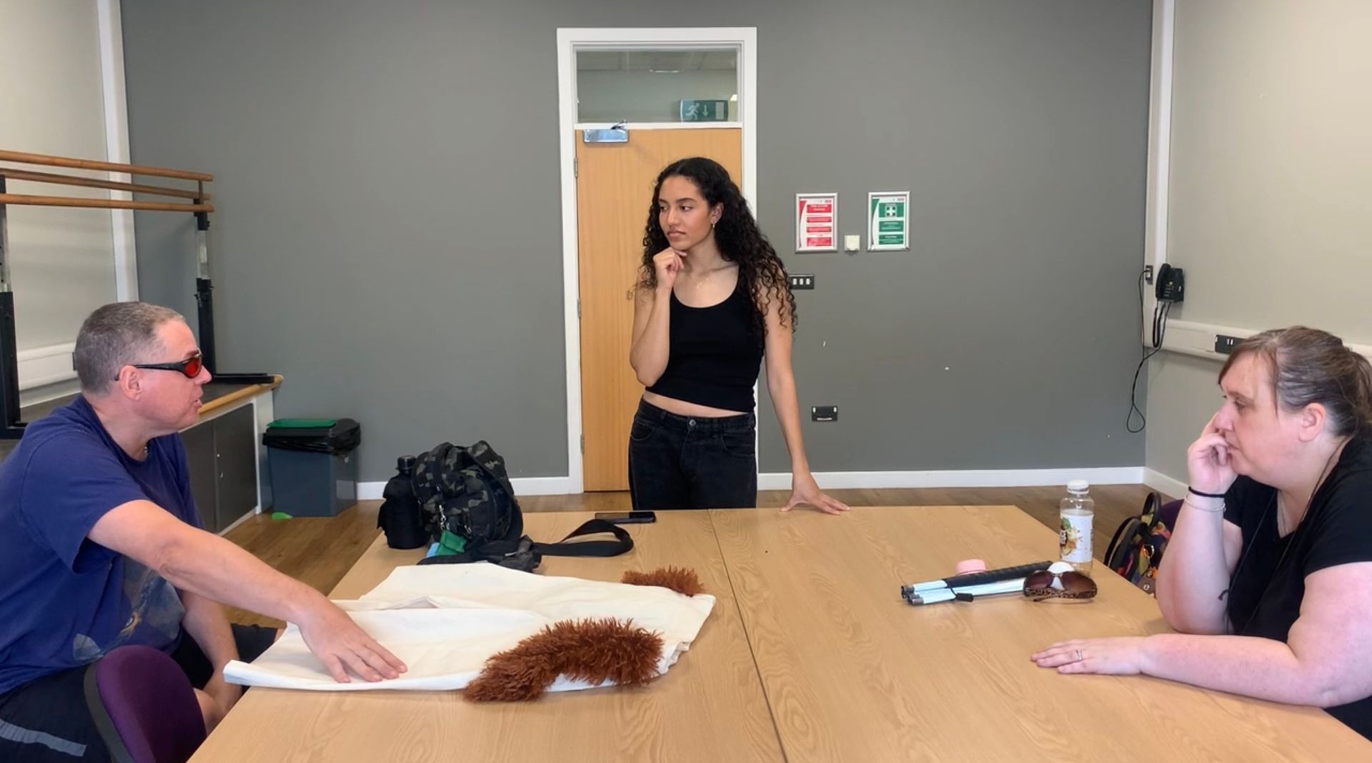 Three people at a table; one points to a fabric, another looks on, a third gestures thoughtfully.