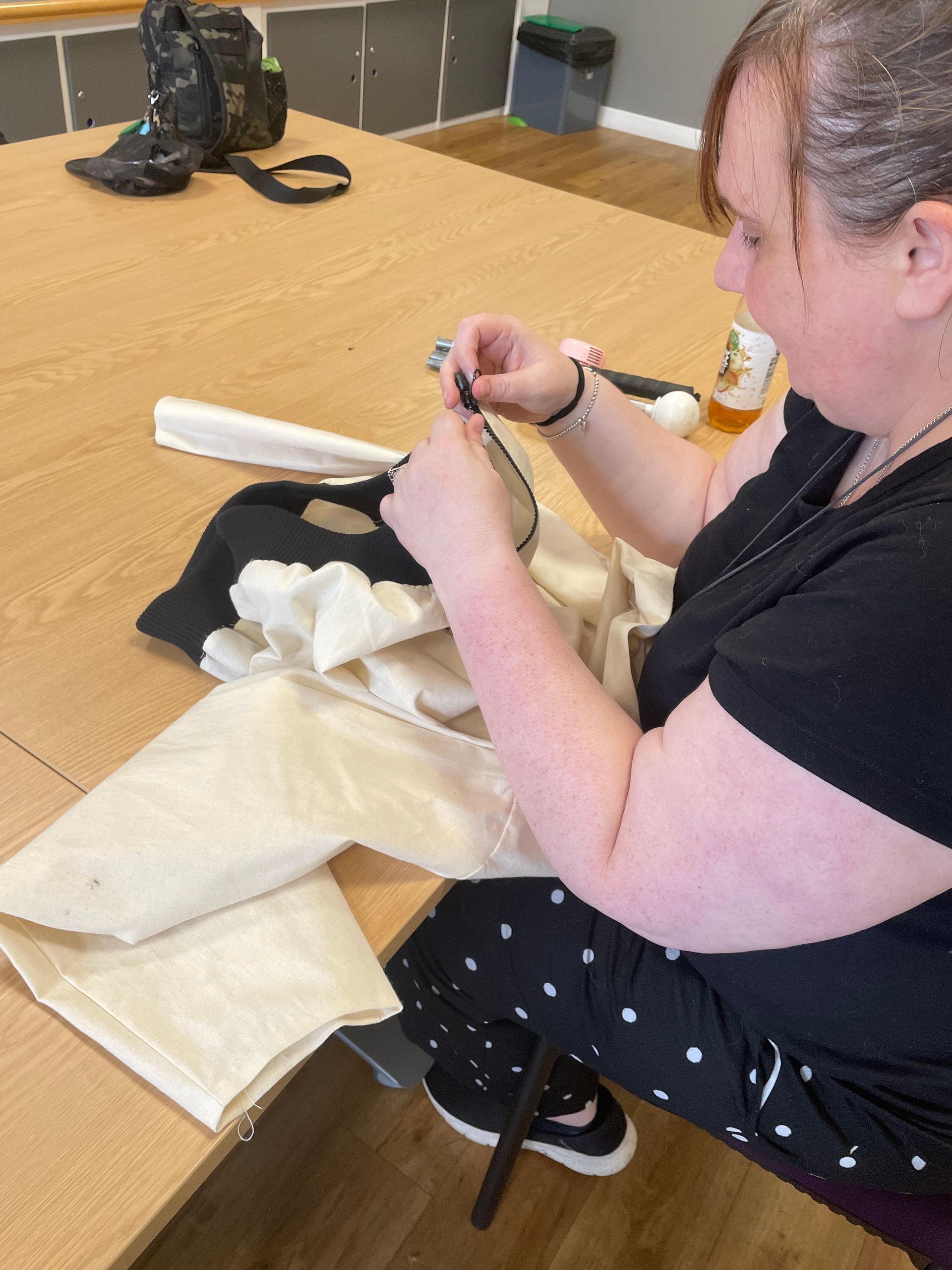 Woman sewing light-colored fabric on a table. She wears a black top, sitting indoors with sewing supplies.
