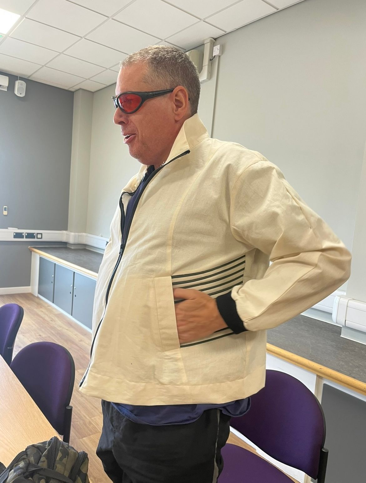 Man in off-white jacket with hand in pocket, red-tinted glasses, in a classroom setting.