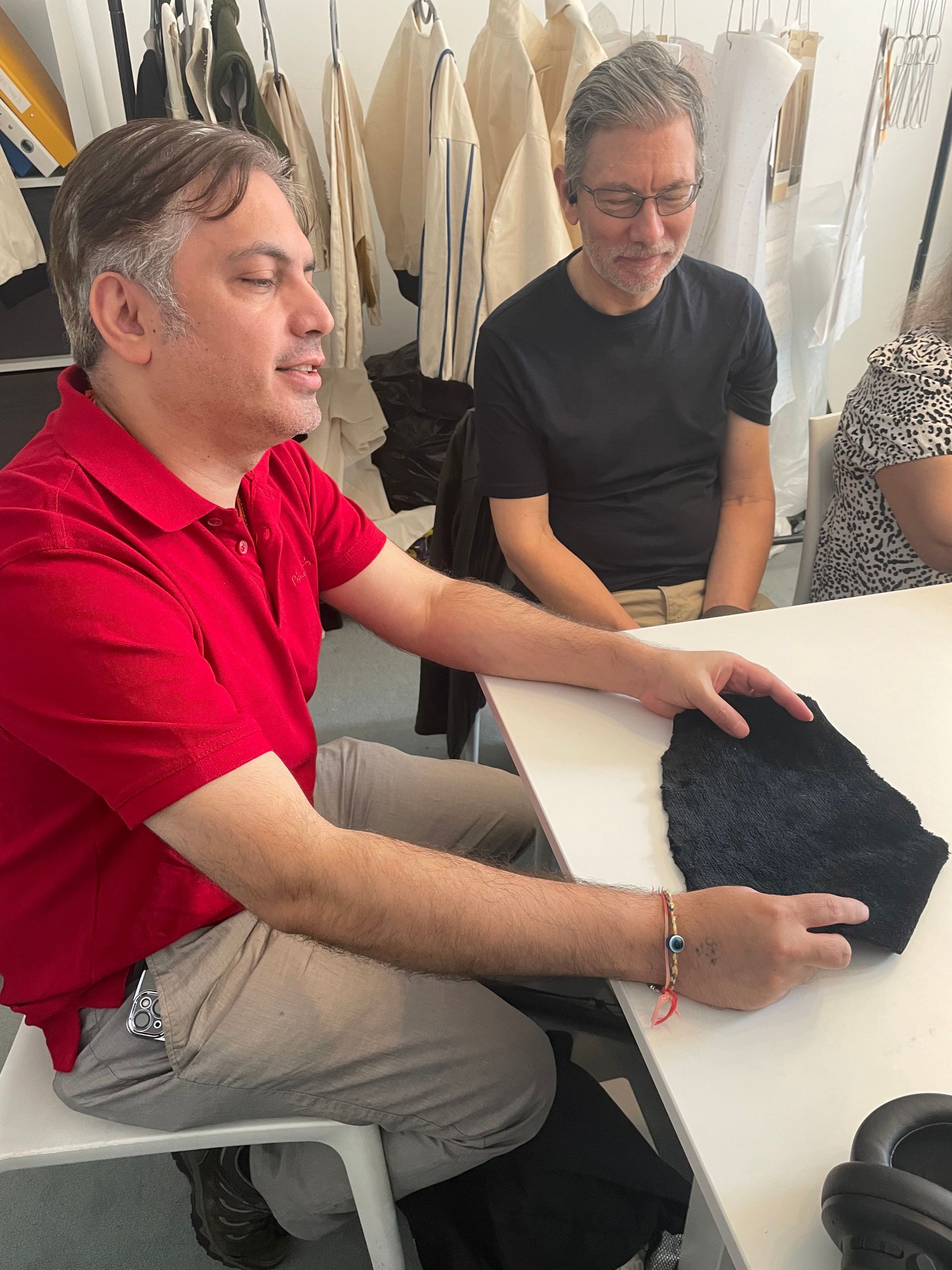 Man in red shirt holding a black fleece piece of material, sitting at a table with two other people in a room with clothing.