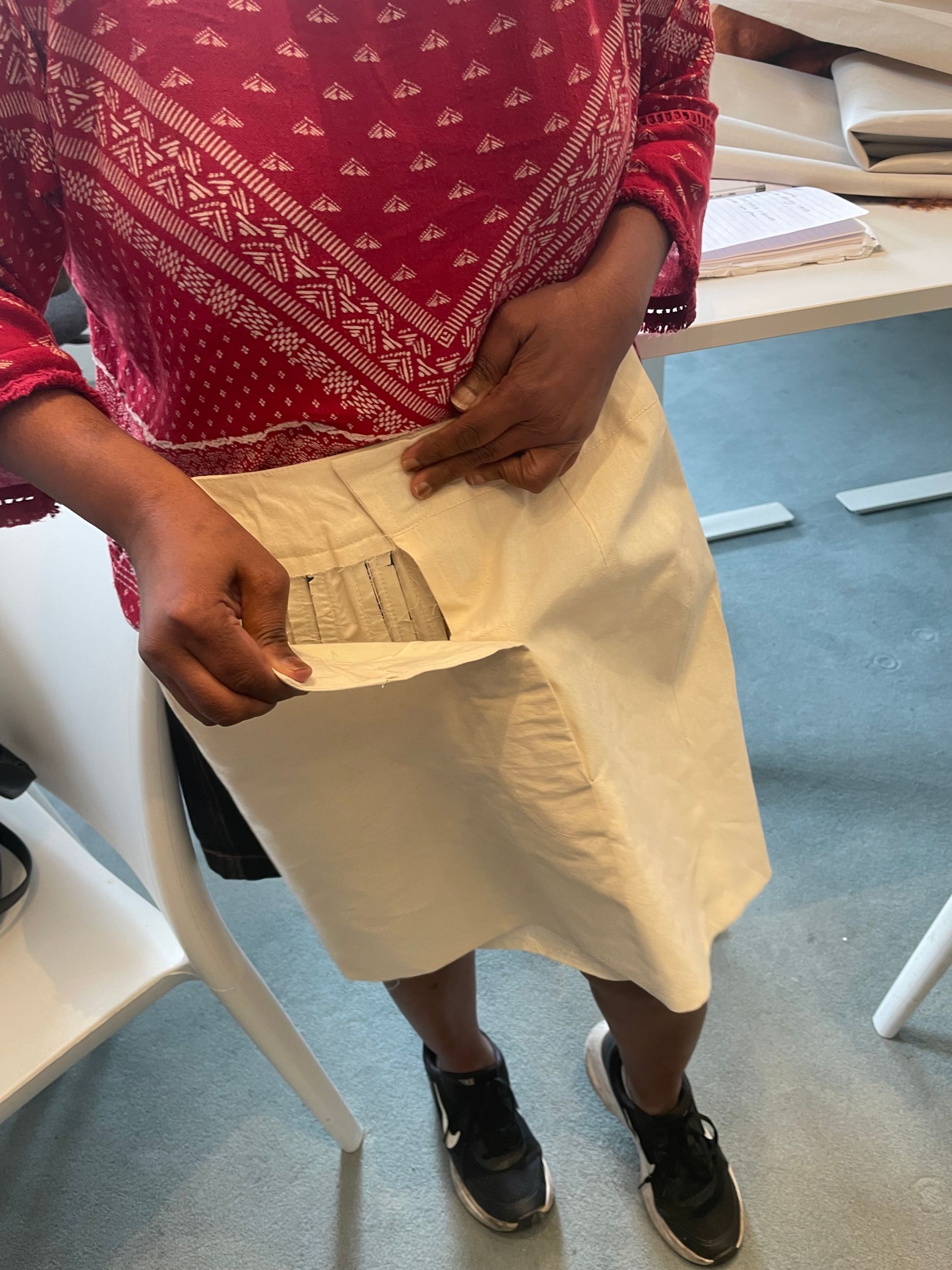 Woman sewing a cream-colored jacket on a wooden table. The jacket has a black zipper. Other items on the table.