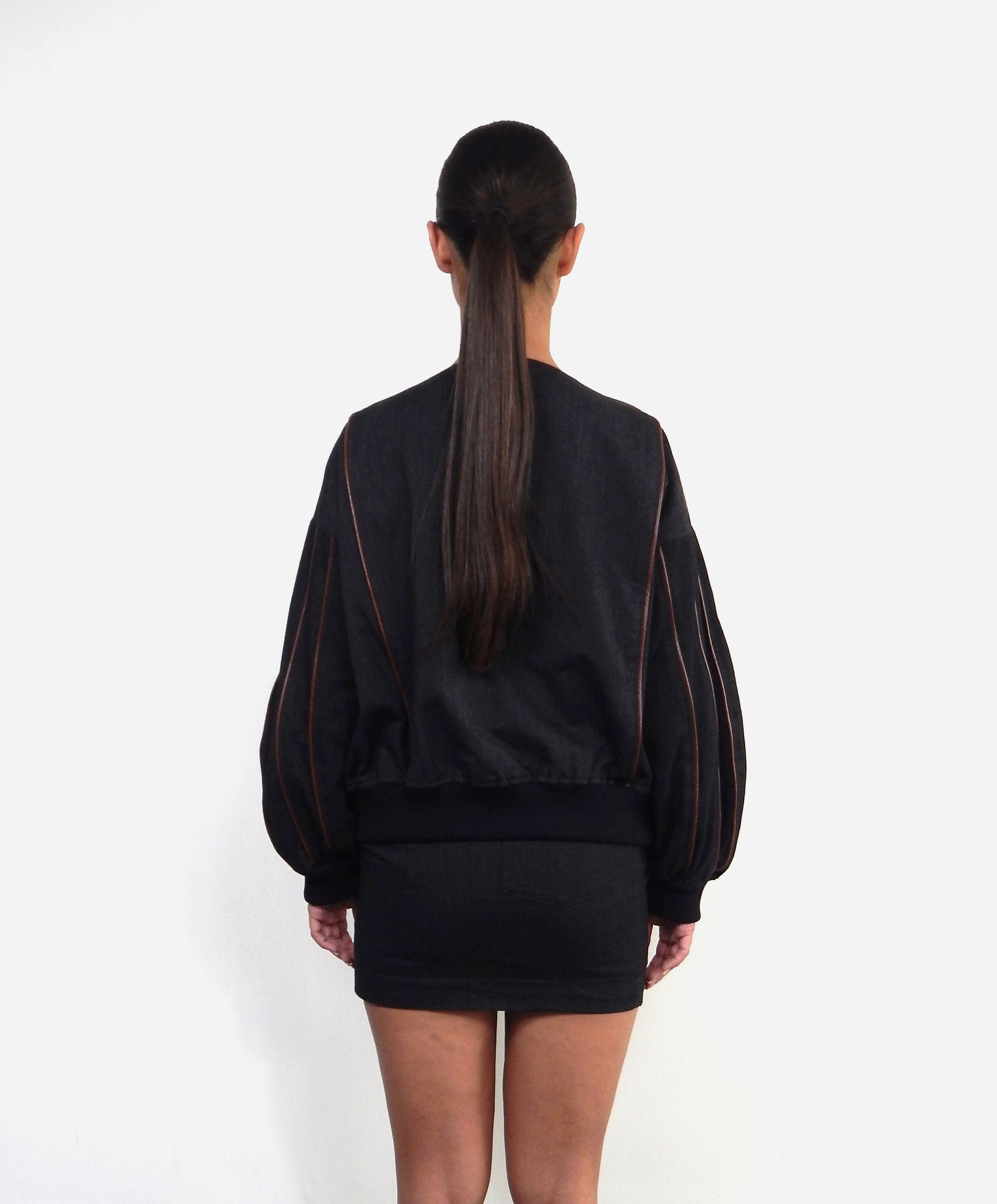 Woman in black bomber jacket and skirt, standing with back to camera, against white background.