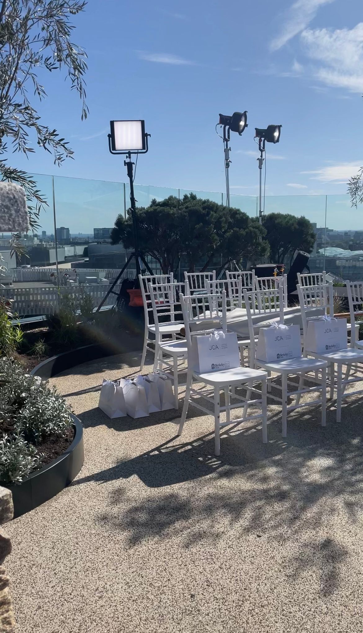 White chairs arranged outdoors, with lighting equipment set up. Sunny day, city view.