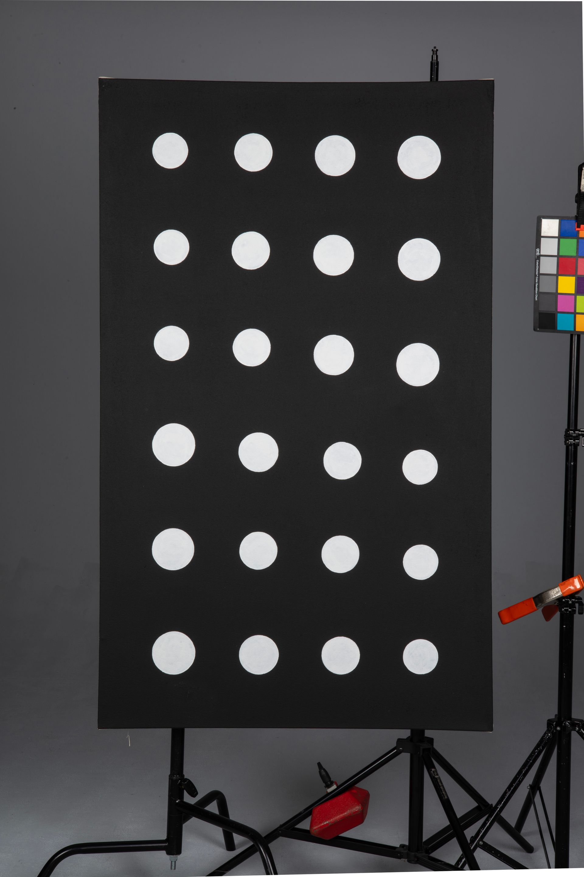 Black panel with 24 white circles in a photo studio, with lighting stands and color calibration chart nearby.