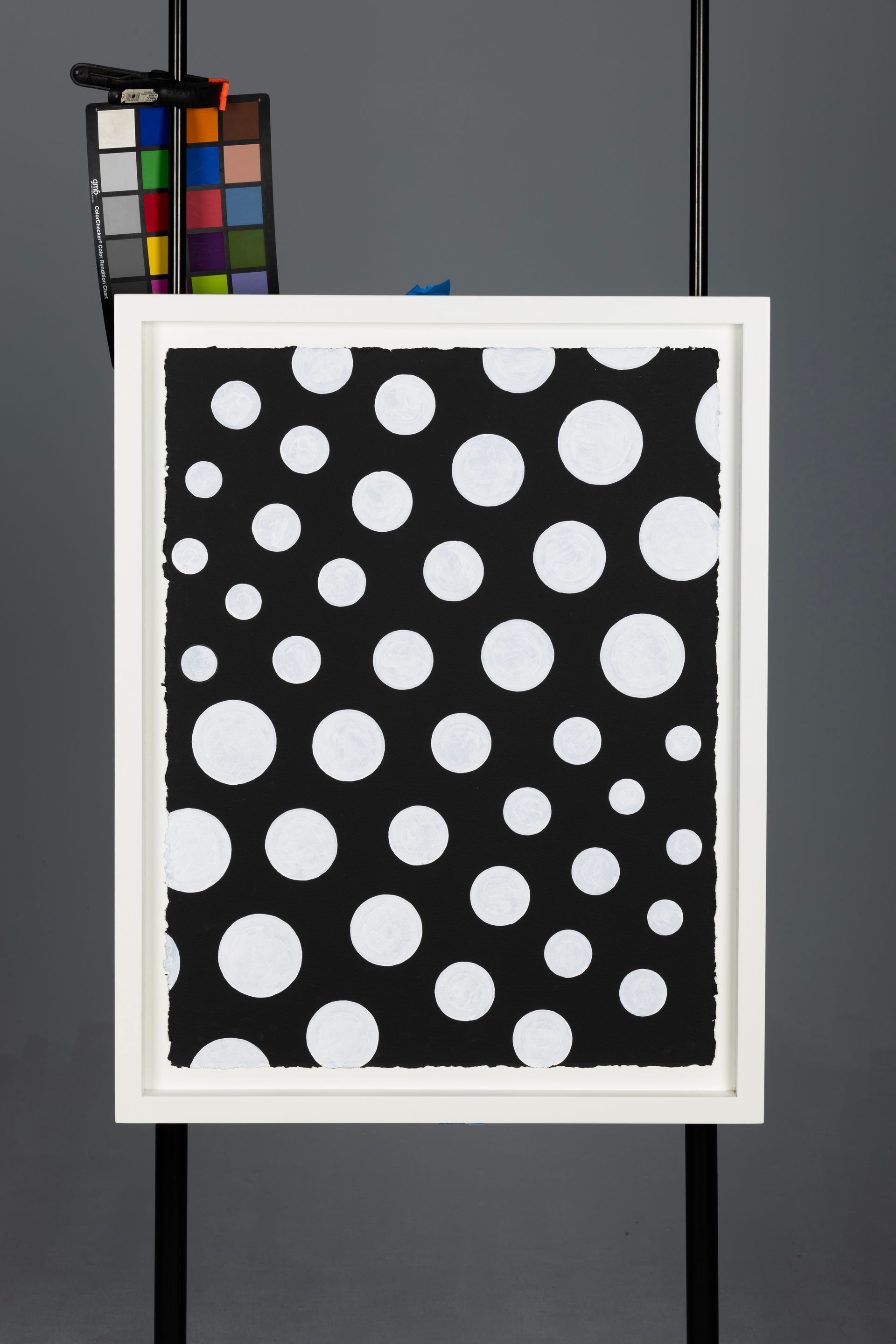 White-framed black panel with scattered white polka dots, hanging on a gray background with color swatches peeking behind.