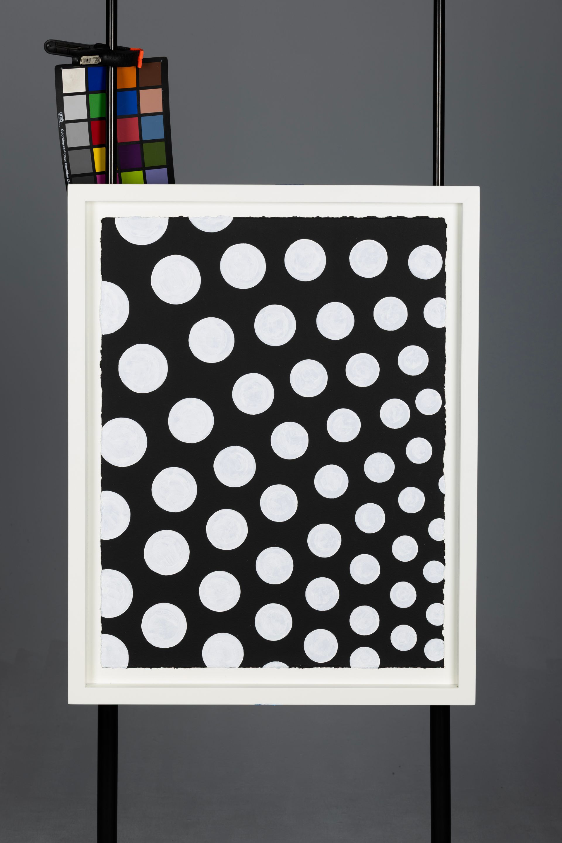 White-framed black artwork with white polka dots, hanging against a gray wall.