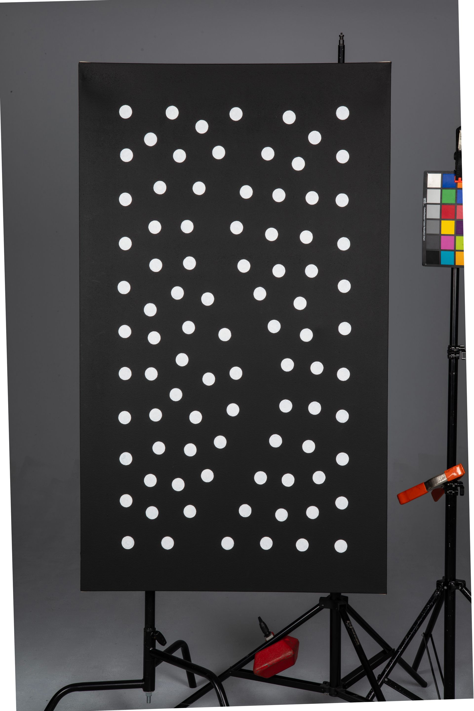 Black panel with rows of white circular markers in a studio, with tripod and color chart beside it.