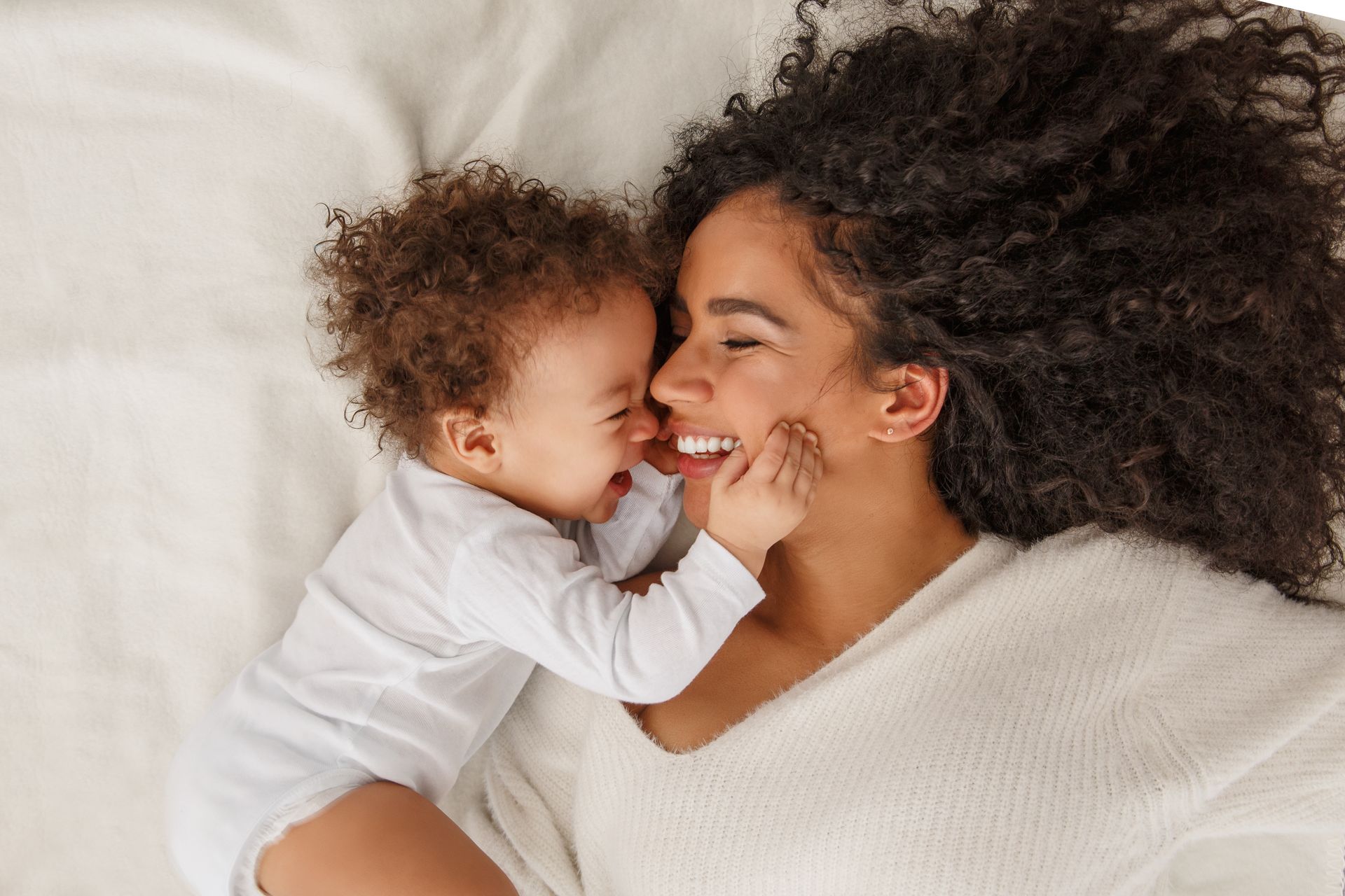 Mother and baby lying on white sheets, laughing and touching faces.