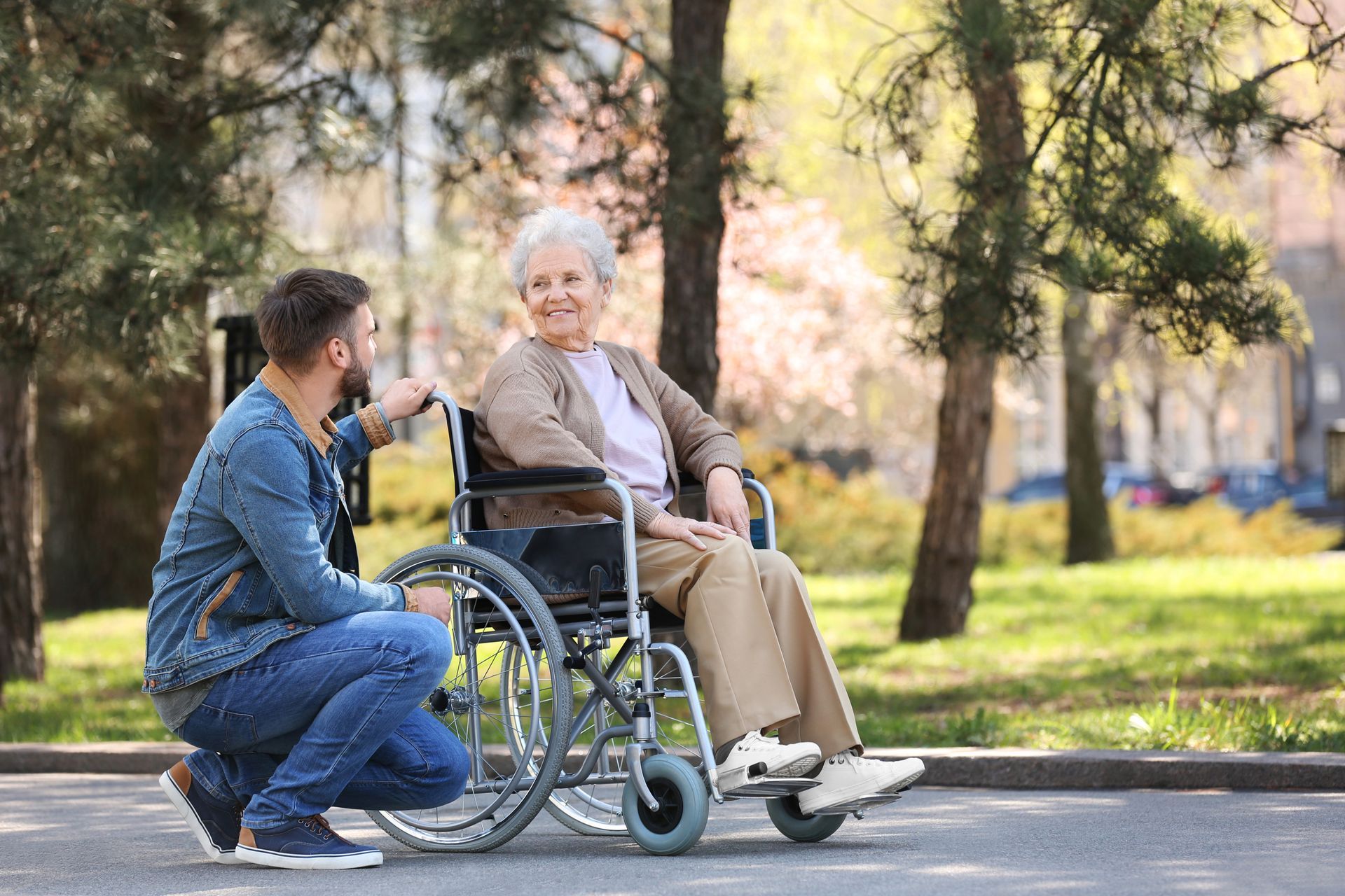Man kneels beside a woman in a wheelchair, outdoors, talking. Both smile.