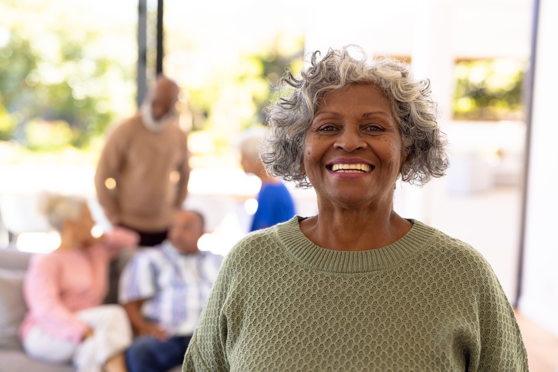 Smiling elderly Black woman in green sweater, other seniors blurred in background.